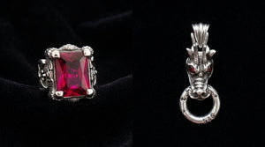 Ruby-red dragon-claw silver ring beside a red-garnet-eyed silver dragon-head pendant on dark velvet.