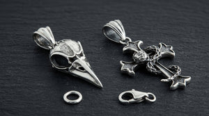 Snake cross and raven skull silver pendants beside a loose end ring and lobster clasp on dark slate.