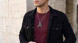 Large sculpted silver scorpion pendant over a burgundy shirt with a black denim jacket beside a stone wall.