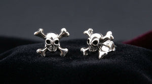 Two skull-and-crossbones silver stud earrings arranged on black cloth.