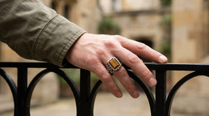 Adult hand in an olive jacket cuff wearing a rectangular tiger’s eye silver ring on a black courtyard railing.
