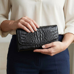 Mujer sosteniendo la billetera tríptico cerrada de cocodrilo hornback negro a la altura de la cintura.