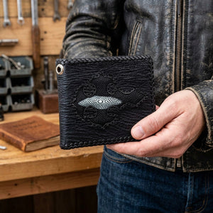 Black shark-skin bifold held beside a leather biker jacket, showing its plain folded spine, three braided edges and pearl-striped stingray cross.