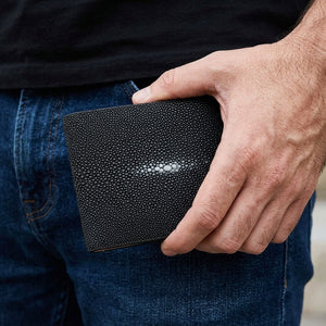 Cartera bifold de piel de raya pulida negra, sostenida cerrada a la altura de la cadera con la parte frontal decorada visible