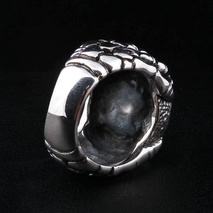 Anillo de plata esterlina con garra esquelética y ojo azul, vista trasera de tres cuartos, el engaste de plata abierto por detrás hacia un hueco oscuro, placas escamadas