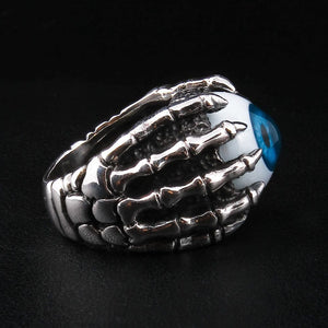 Anillo de plata esterlina con garra esquelética y ojo azul, ángulo de tres cuartos sobre superficie oscura, garras de plata esterlina agarrando el ojo, su iris azul