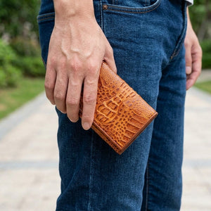 Cartera tríptico cuadrada marrón de piel de lomo de cocodrilo sostenida junto a tela vaquera oscura