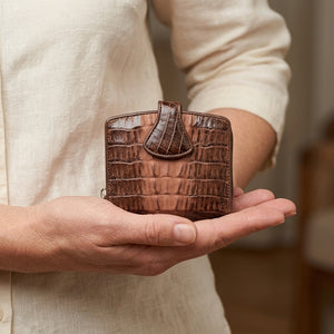 Billetera con broche de piel de cola de cocodrilo marrón bicolor sostenida por la mano de una mujer en una vista de cintura de cerca, mostrando su lengüeta oscura en forma de lágrima