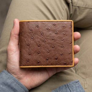 Brown quill ostrich bifold with tan edging supported in a hand above a khaki-clothed lap.