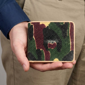 A hand holds the camouflage stingray bifold with tan trim and its pale pearl cluster visible.