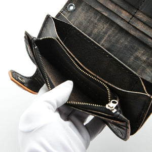 Portefeuille long en cuir noir à motif tête de mort et os sculptés, compartiment zippé maintenu ouvert par une main gantée