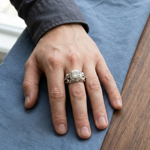 Clear round topaz and fleur-de-lis silver ring on a hand resting on blue cotton beside dark oak.