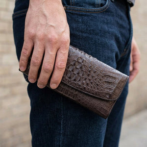 Billetera trifold de cocodrilo hornback color cacao sostenida junto a tela vaquera oscura