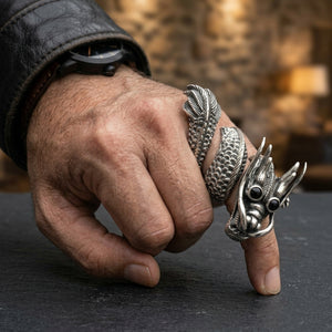 Anillo de plata con dragón enroscado y ojos de circonita cúbica negra en una mano flexionada junto a una mesa de pizarra oscura.