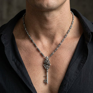 Sterling zilveren halsketting met kroonsleutel gedragen bij een open donkere halslijn, met weergave van de decoratieve ketting, hals en volledige sleutelhanger.