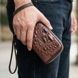 Cartera con doble cremallera de lomo de cocodrilo marrón oscuro sostenida junto a tela vaquera oscura