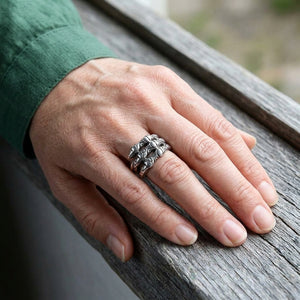 Der gravierte Krallenring umschließt einen Finger an einer Hand, die neben einer grünen Manschette auf Holz ruht.