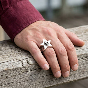 Una mano adulta che indossa l'anello a stella con teschio riposa su legno invecchiato accanto a un polsino bordeaux.