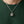 Een kleine zilveren kattenhanger met goudkleurige florale ogen en een voorhoofdskruis hangt aan een stylingketting over een donkergroen overhemd.
