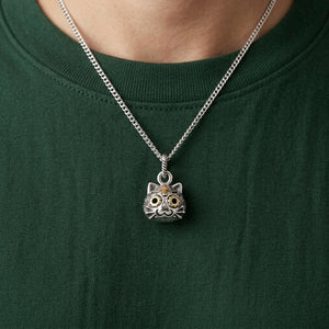 Een kleine zilveren kattenhanger met goudkleurige florale ogen en een voorhoofdskruis hangt aan een stylingketting over een donkergroen overhemd.