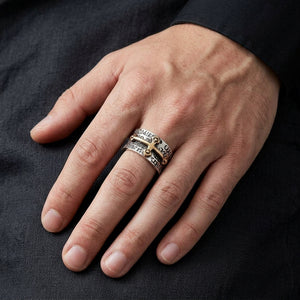 Hand resting on dark fabric, wearing the wide sterling silver ring with a raised gold-plated cross and recessed lettering.