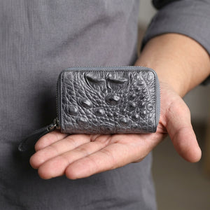Cartera de cocodrilo gris oscuro con doble cremallera sostenida por la mano de un hombre en vista cercana de cintura, con correa de muñeca y textura dorsal visibles