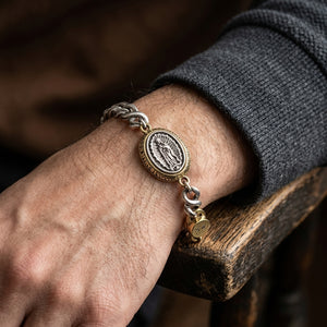 Silver-and-brass Guadalupe medallion bracelet worn on a wrist beside a charcoal sleeve.