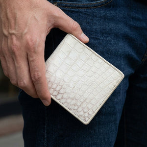 Cartera bifold de cuero de vientre de cocodrilo en color marfil sostenida junto a tela vaquera oscura