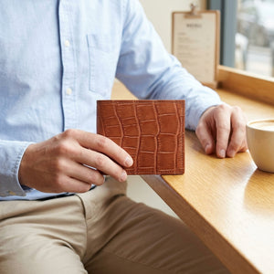 Light-brown crocodile belly bifold held beside a pale-blue shirt at a wooden cafe table, showing rectangular scales and stitched edges.