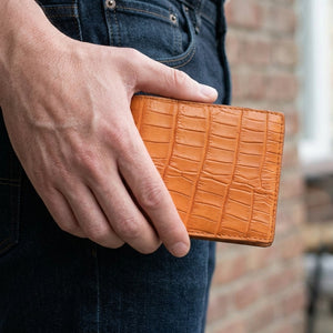 Billetera bifold de piel de vientre de cocodrilo marrón claro sostenida junto a tela vaquera oscura