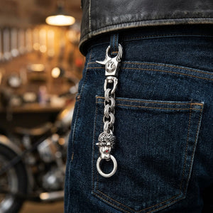 Close-up of a lion-head silver keychain with flame-engraved links and red eye details, clipped through a dark jeans belt loop beside the back pocket.