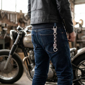 Red-eye silver lion keychain clipped through a denim belt loop beside a black leather biker jacket, with three flame links and a split key ring above the back-pocket tip.