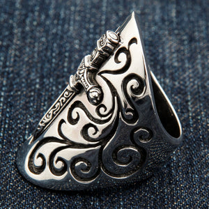 Three-quarter close-up on denim, the dagger raised down the center of a sterling silver shield cut with scrollwork.