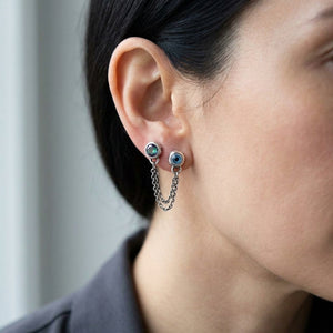 Mystic topaz and blue eye silver studs connected by two draping chains across two exposed earlobe piercings in a natural side profile.