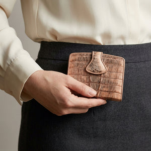 Cartera con broche de presión de cola de cocodrilo en rosa viejo sostenida a la cintura de una mujer, con lengüeta en forma de lágrima y escamas de piel bitonales visibles