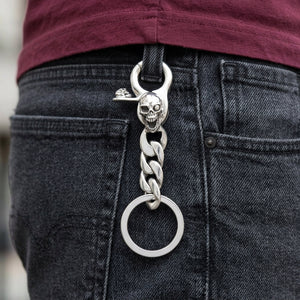 Nøkkelring med sølvhodeskalle med åpen kjeve festet i beltestroppen på en koksgrå bukse, med panserlenker og splittringen hengende nedenfor.