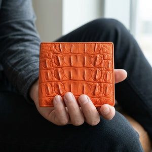 Cartera bifold de piel de lomo de cocodrilo naranja sostenida naturalmente en una mano adulta