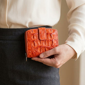 Cartera de piel de cocodrilo hornback naranja con broche sostenida cerca de la cintura de una mujer, mostrando las escamas de piel en relieve y la presilla con broche