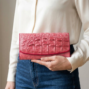 Cartera larga trifold de piel de lomo de cocodrilo rosa sostenida naturalmente por una mujer adulta