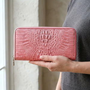 Cartera larga de piel de cocodrilo hornback rosa sostenida en una mano adulta