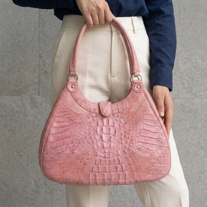 Borsa a spalla in pelle di coccodrillo hornback rosa tenuta per i manici all'altezza dei fianchi con camicia blu navy e pantaloni crema.