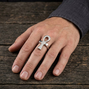 Anello ankh in argento lucidato indossato sulla mano di un adulto appoggiata su legno scuro accanto a una manica grigio antracite.