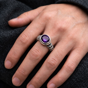 Anillo de plata con amatista morada y diseño de columna usado en una mano con una piedra violeta más profunda y reflejos de facetas visibles