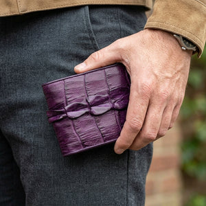 Lilla krokodillehale bifold-lommebok holdt ved siden av grå jeans, med den opphøyde haleryggen vendt fremover.