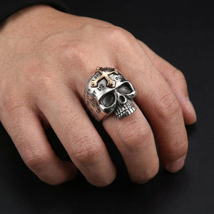 Adult hand wearing the R2586 sterling silver skull ring with a 14K yellow gold cross on a charcoal background