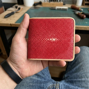 Red stingray bifold with tan leather trim supported in a hand above blue jeans, showing its small pale pearl cluster.