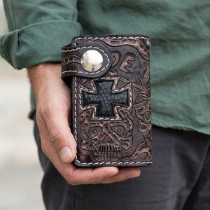 A hand holds the brown tooled Ride to Live wallet with its black lizard cross and skull carving facing forward.