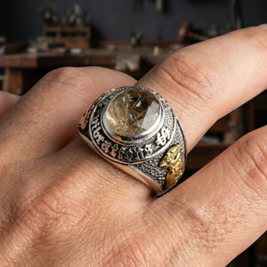 Rutilated quartz tri-metal silver ring worn on a hand, showing the faceted stone and brass money-bag detail against a blurred workshop background