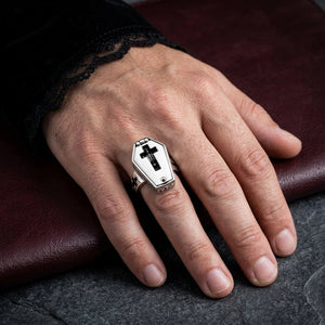 Closed cross-window coffin ring worn on a hand beside a black velvet and lace cuff.