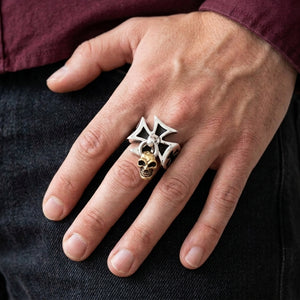 Silver cross ring with a dangling gold-tone skull worn on a relaxed hand beside a burgundy cuff.